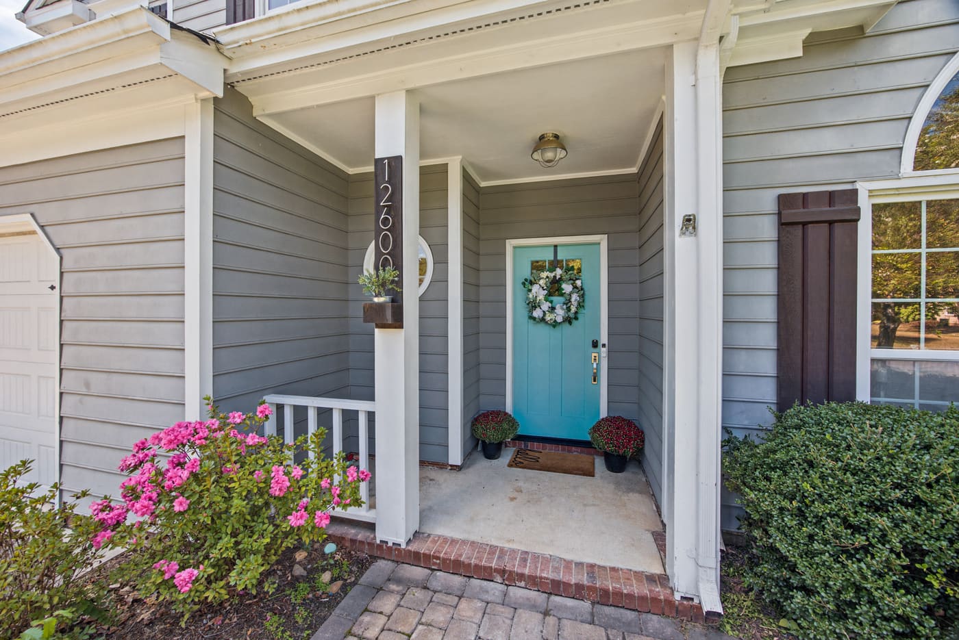 The covered front entry with a turquoise door, a wreath, potted mums and a brick paver walk lined by pink azaleas.